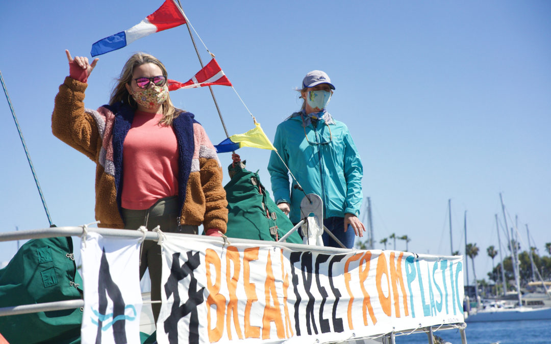 Two women on the bow of a boat behind a banner saying #Break Free From Plastic