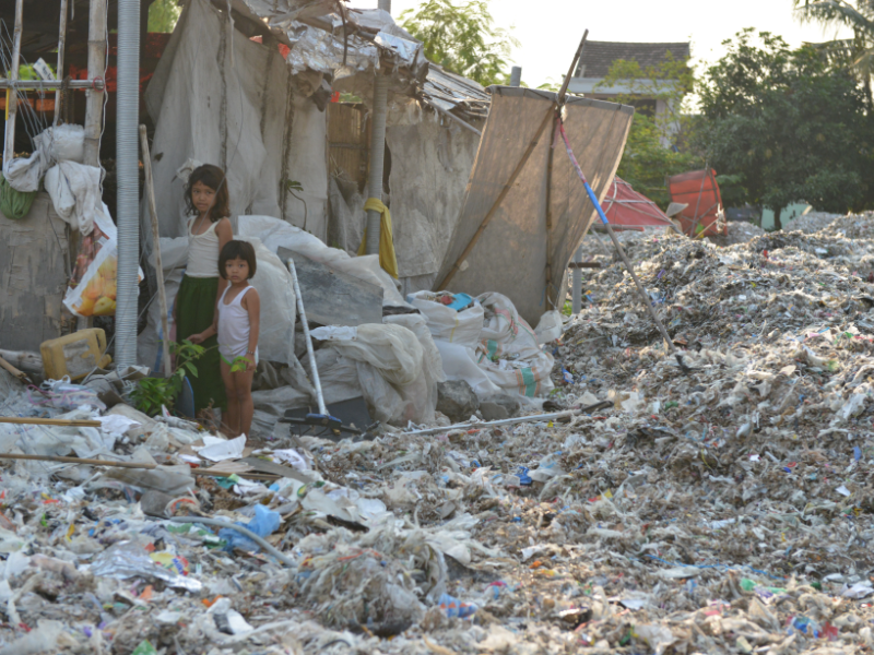 Two girls standing outside of a shack with plastic film waste covering the ground.