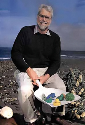 Man, Curtis Ebbesmeyer, sits on beach with found plastic objects.